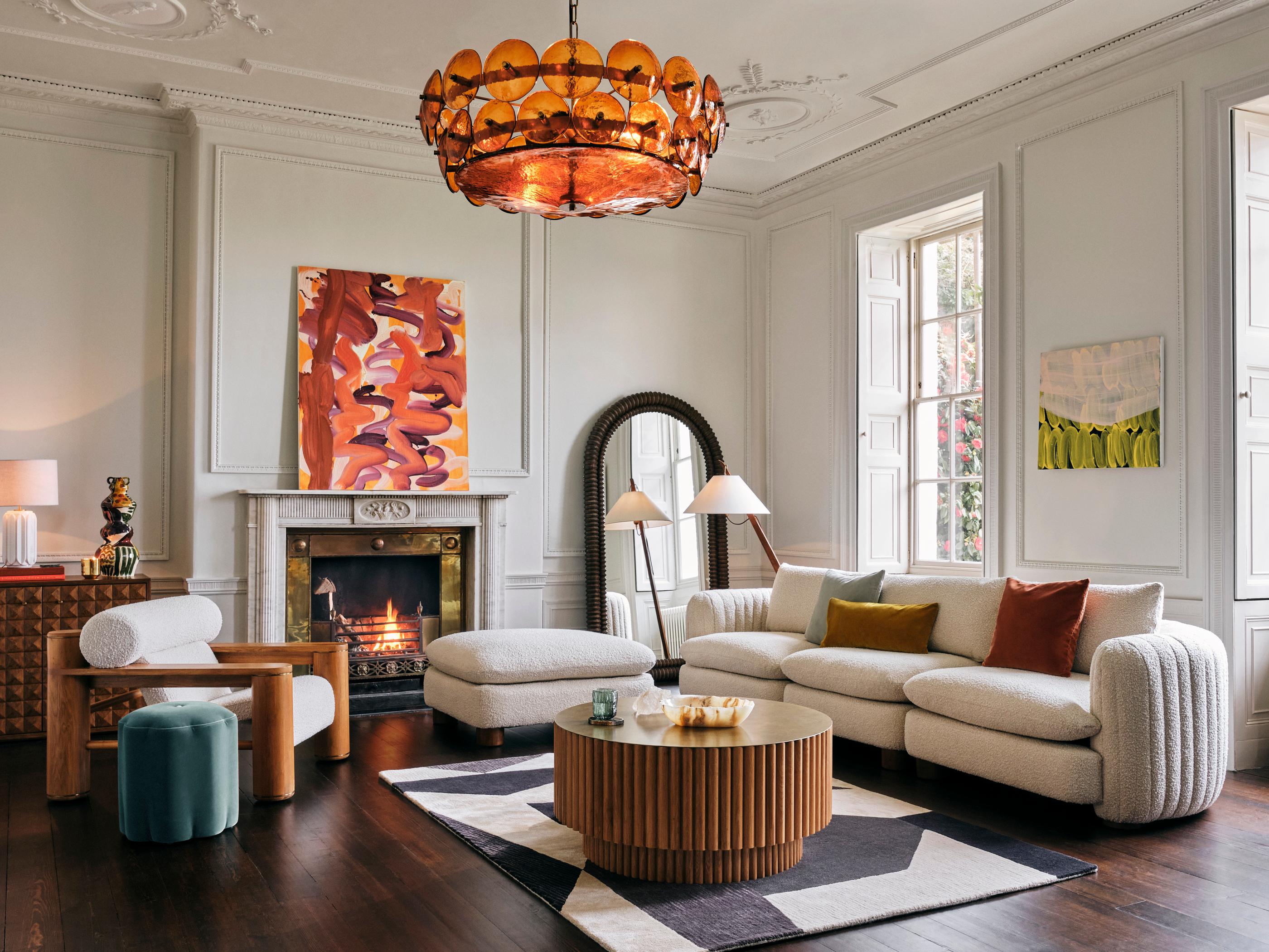 A stylish living room features a comfortable beige sofa and armchair around a wooden coffee table. A fireplace with a vibrant abstract painting above it and large windows illuminate the space.