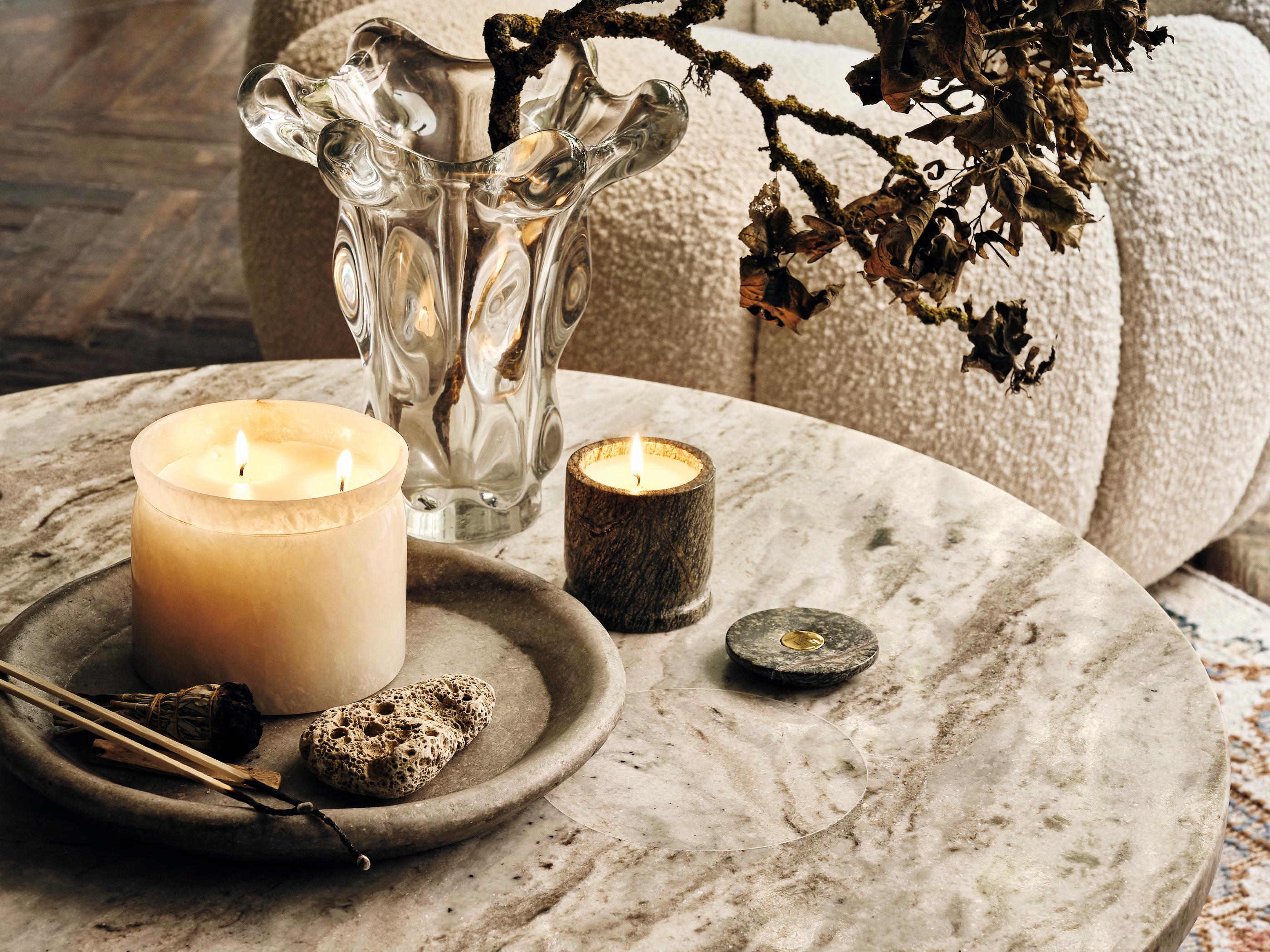Lit candles and dried branches rest on a round marble table, accompanied by a stone tray with incense sticks and a pumice stone, all placed in a cozy, textured living room.