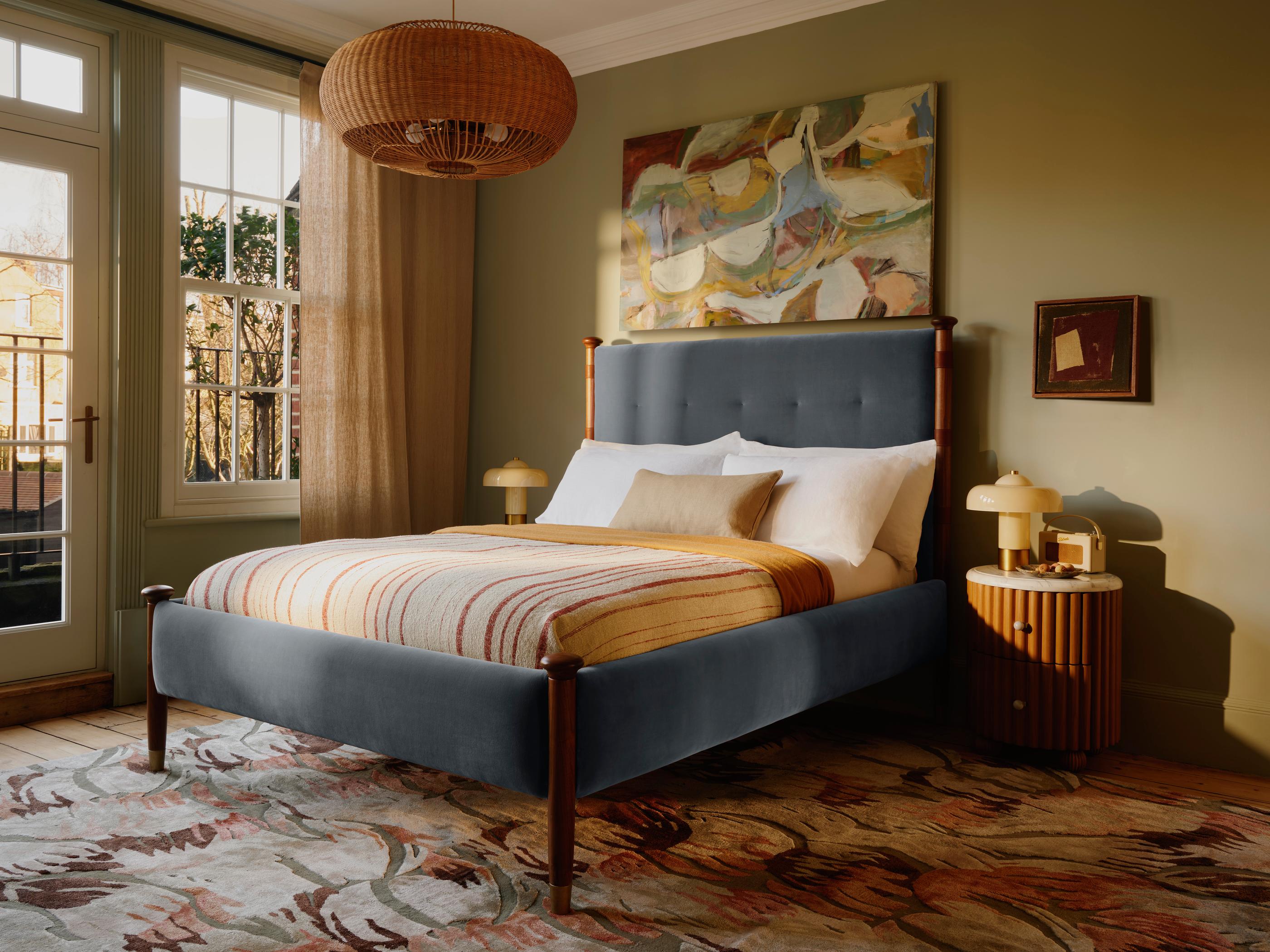 Bed adorned with striped bedding in a warmly lit bedroom. On the nightstand is a lamp, abstract painting above the bed, rug on the wooden floor, and large windows with beige curtains.
