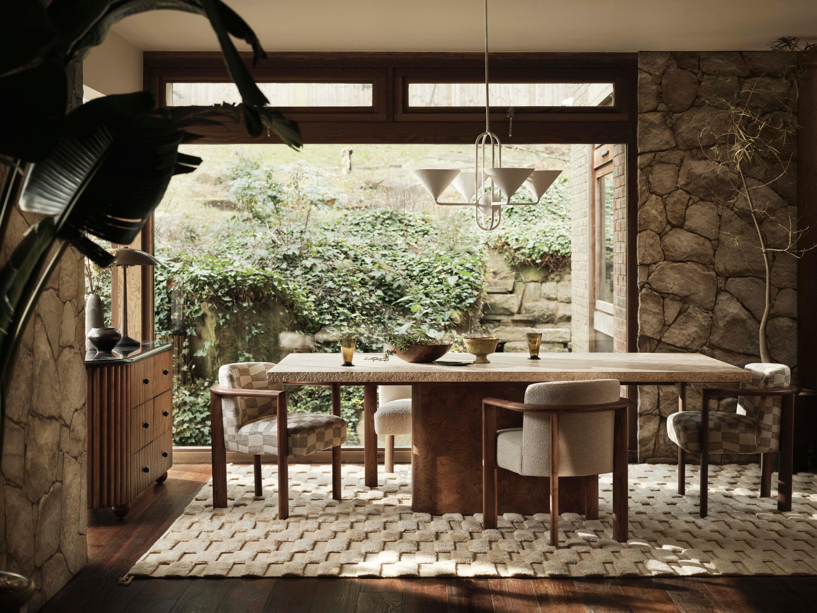 A dining table surrounded by cushioned chairs with a pendant light overhead in a stone-walled room, featuring large windows, green foliage outside, a patterned rug, and wooden furniture.
