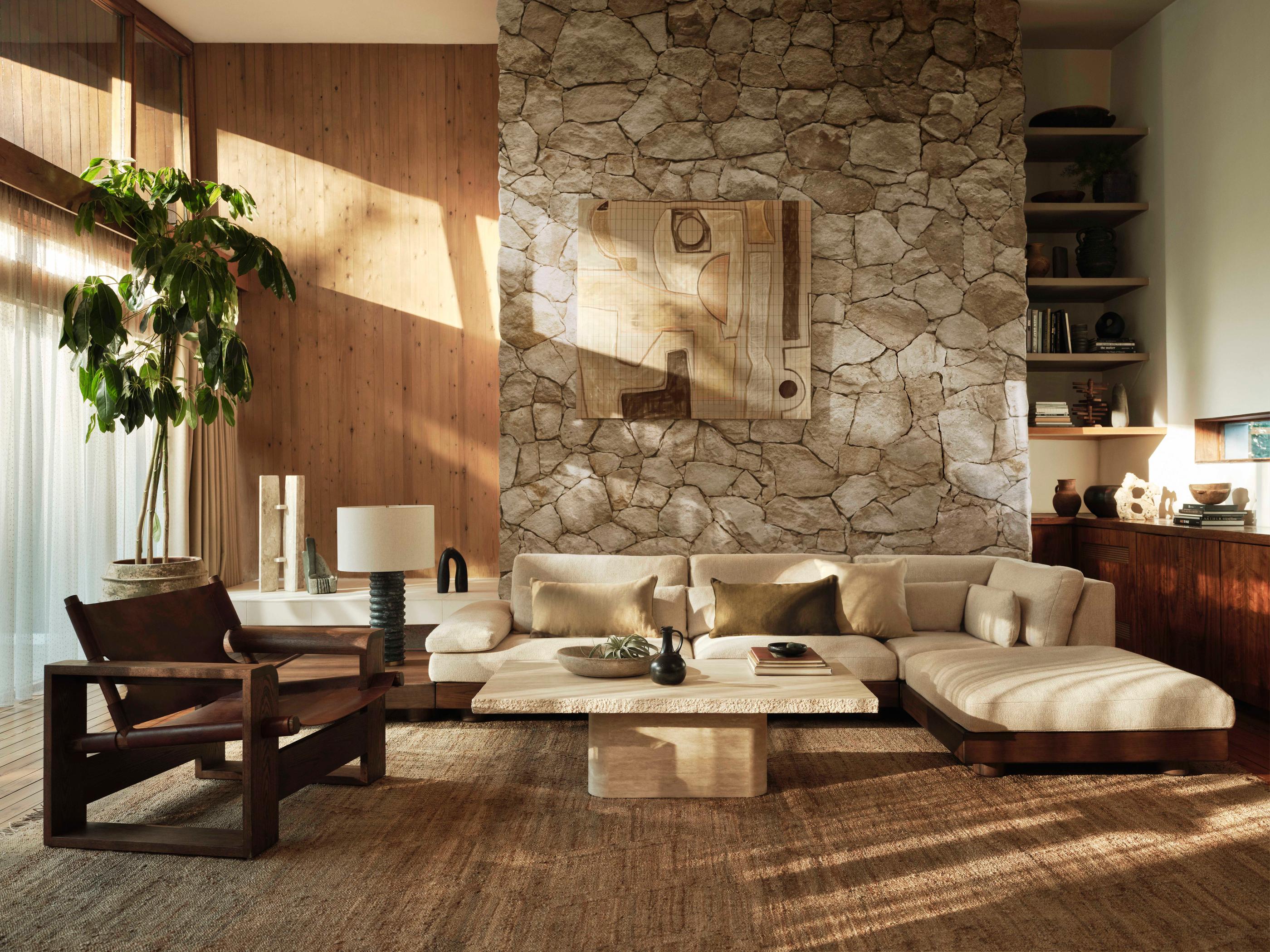 A beige sectional sofa sits in a warm, sunlit living room with stone and wood-paneled walls, featuring a large potted plant, abstract wall art, wooden chair, and shelves with books and decor.