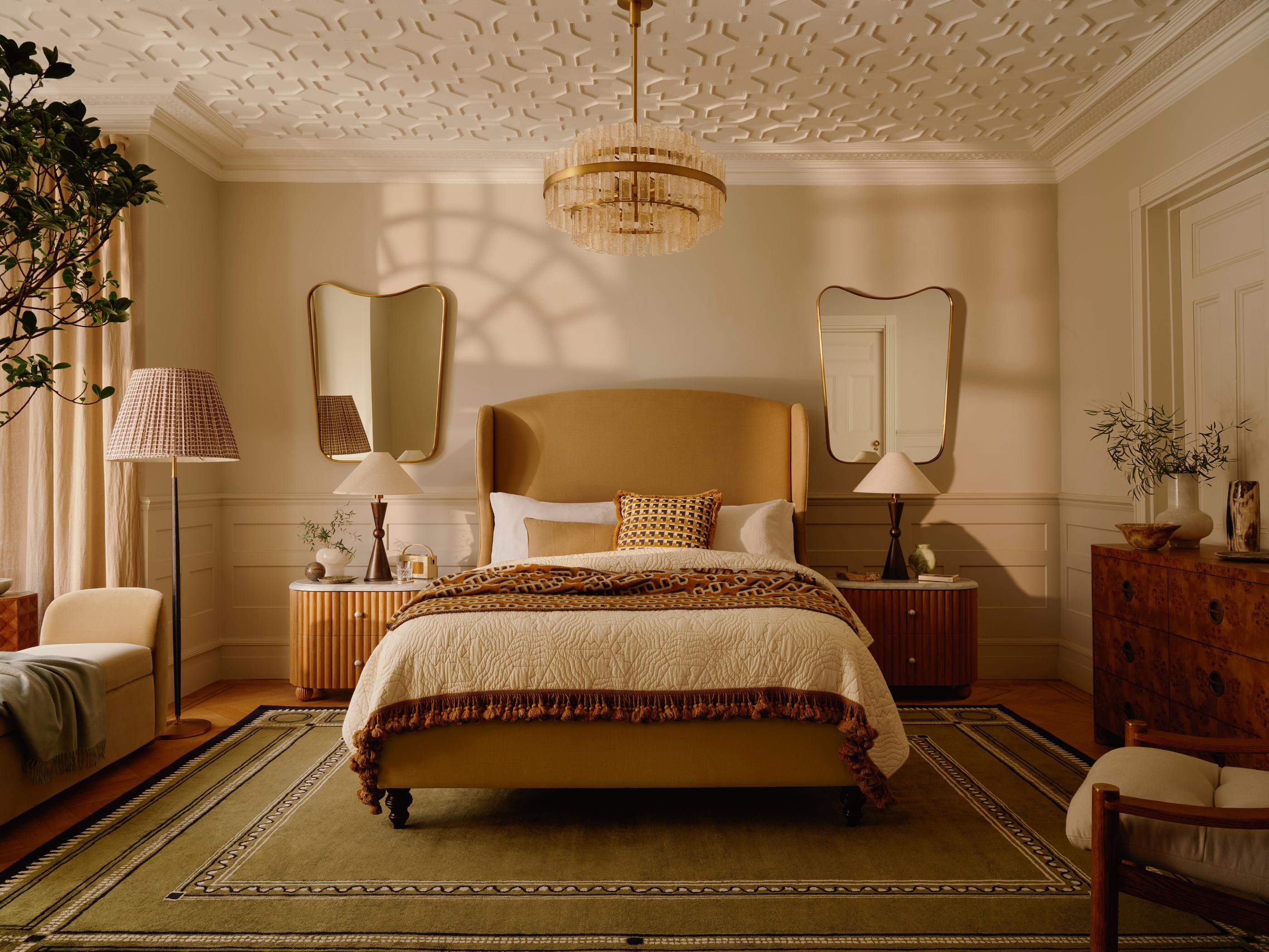 A bed with a beige headboard and decorative pillows is situated in a stylish bedroom, featuring matching nightstands, mirrored walls, a chandelier, an armchair, and a textured ceiling.