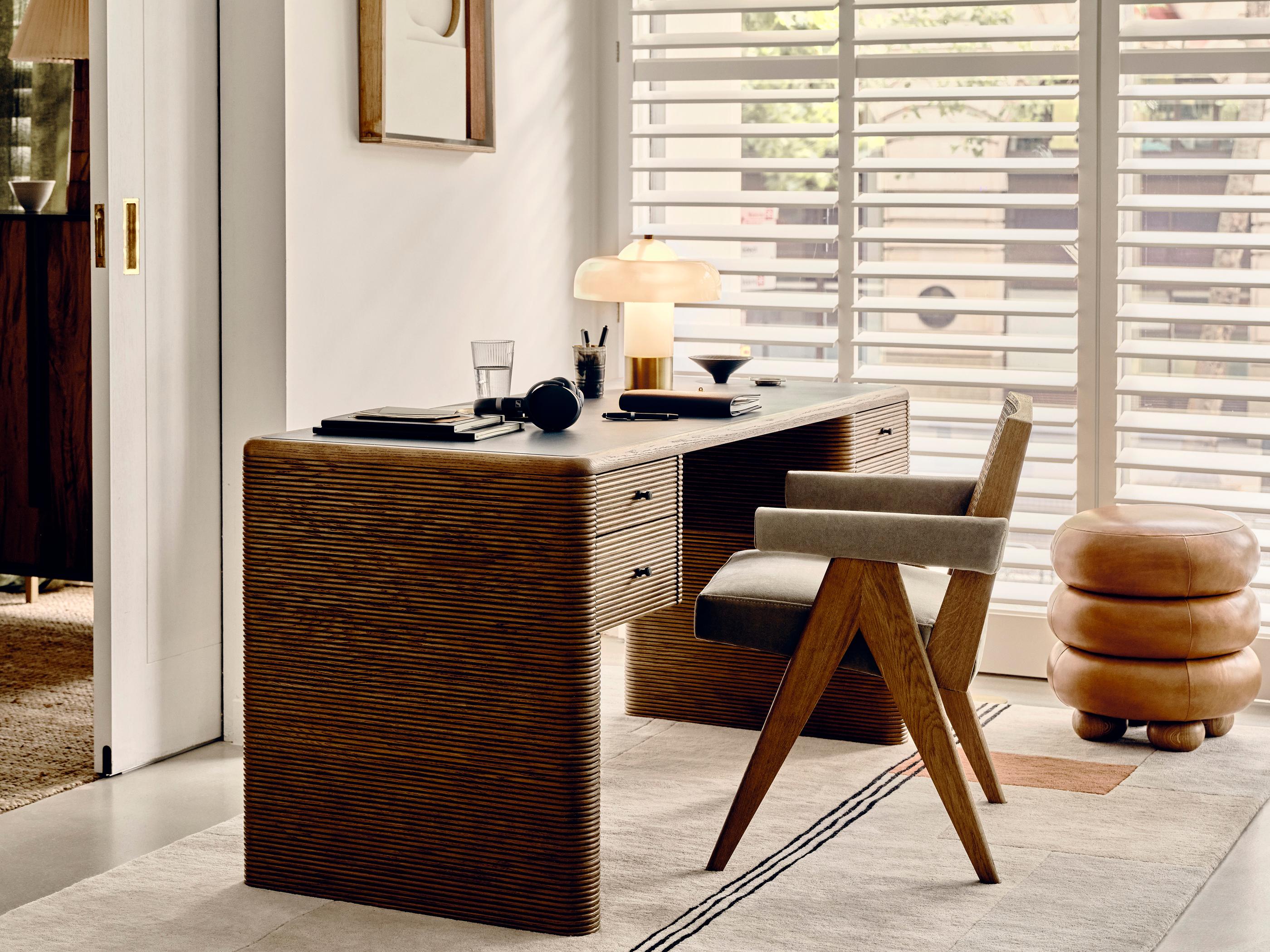 A modern wooden desk with drawers holds books, a lamp, and stationery. A cushioned chair sits at the desk. The desk is positioned against a wall with a framed painting.