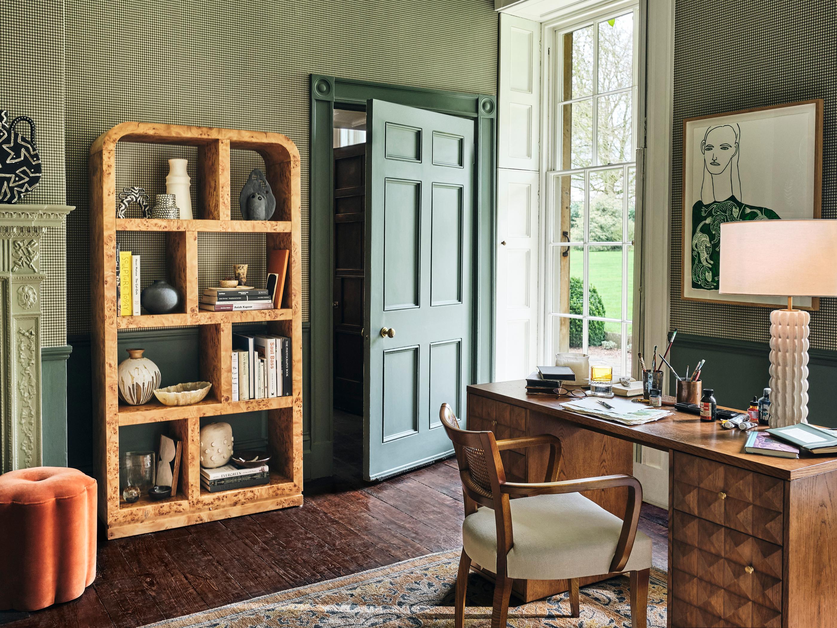 A cozy study room featuring a wooden desk with a lamp, chair, and art supplies, adjacent to a tall wooden bookshelf filled with decor and books, near a large window overlooking greenery.