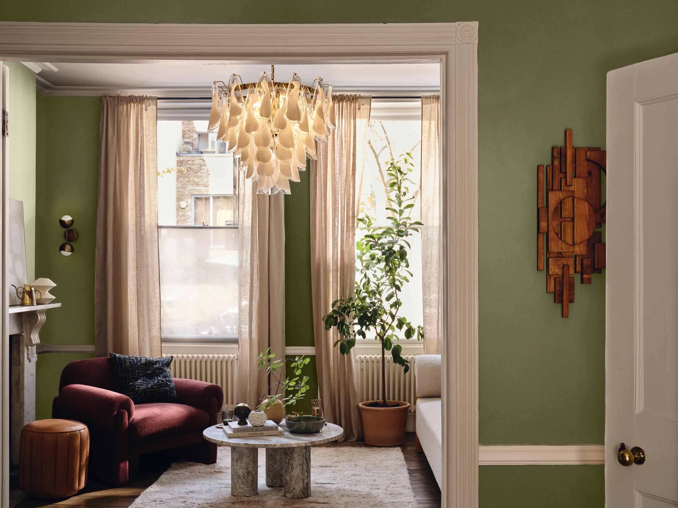A red chair sits beside a round marble coffee table with plants, in a green-walled living room featuring large windows with beige curtains, an elaborate chandelier, and decorative wooden wall art.