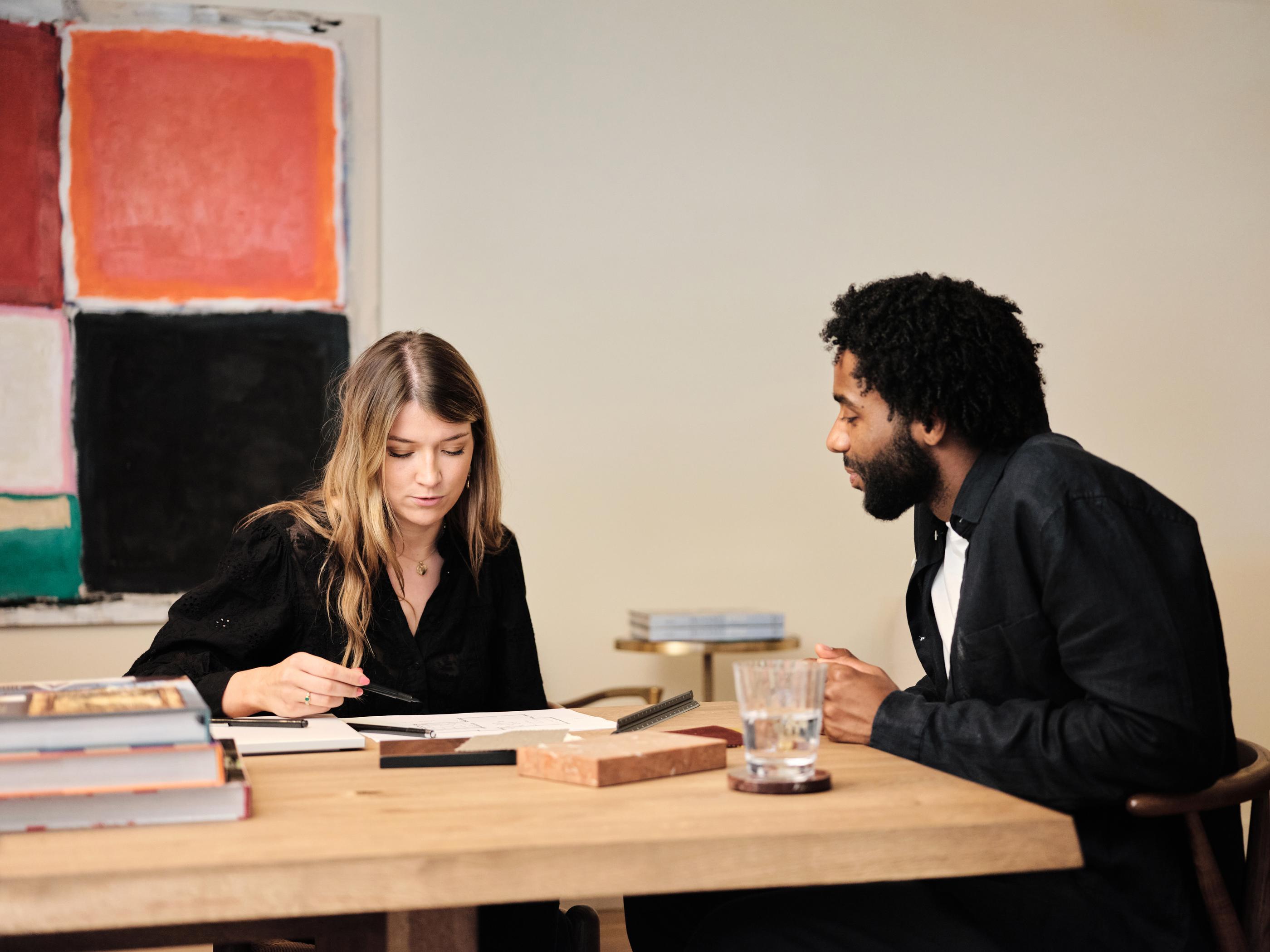 Two people sit at a wooden table discussing documents, surrounded by books, with a large abstract painting in the background.