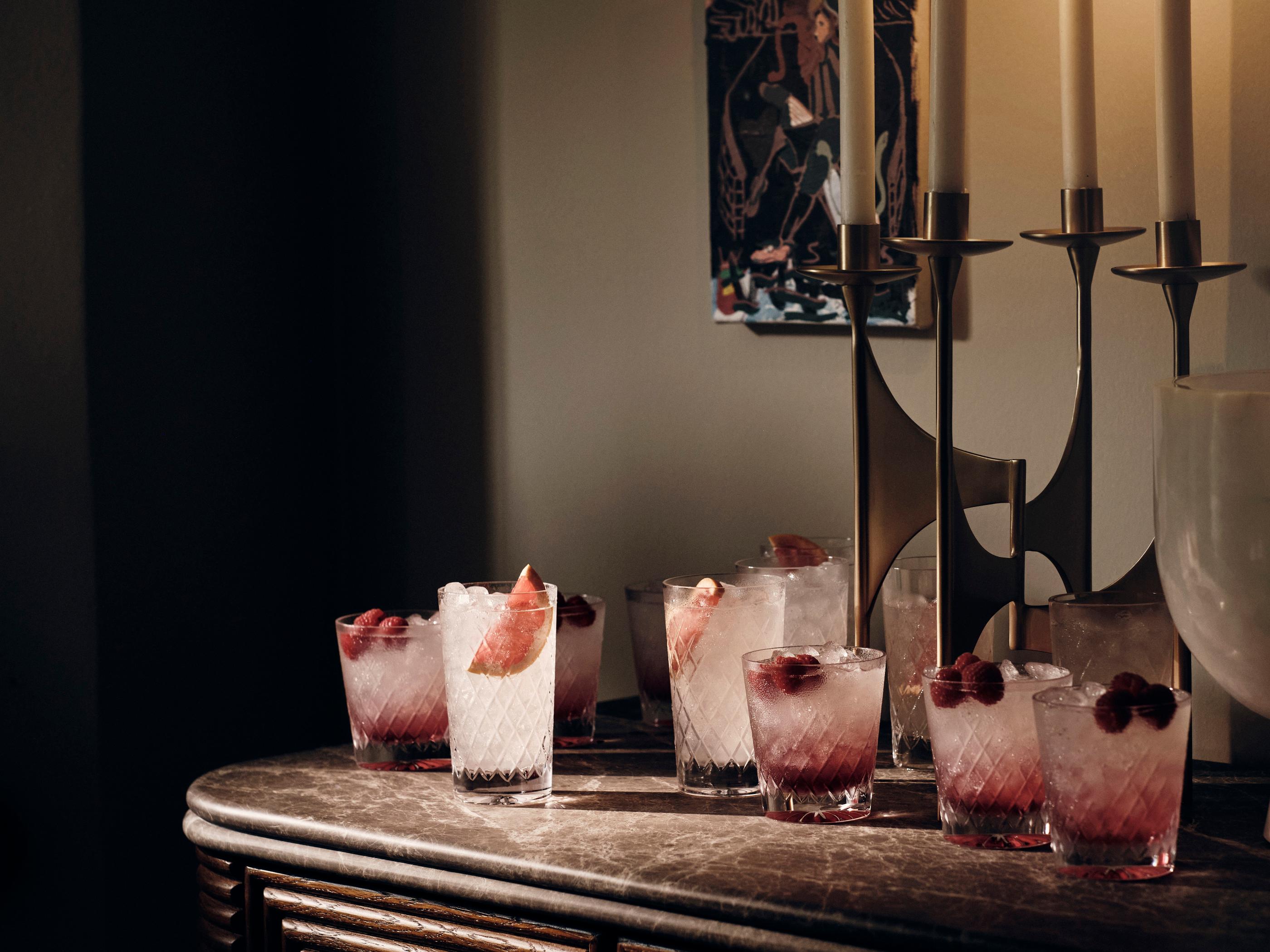 Several glasses filled with ice, raspberries, and a pink liquid rest on a marble-topped table. Behind them, a brass candelabra with white candles and a framed artwork adorn the wall.