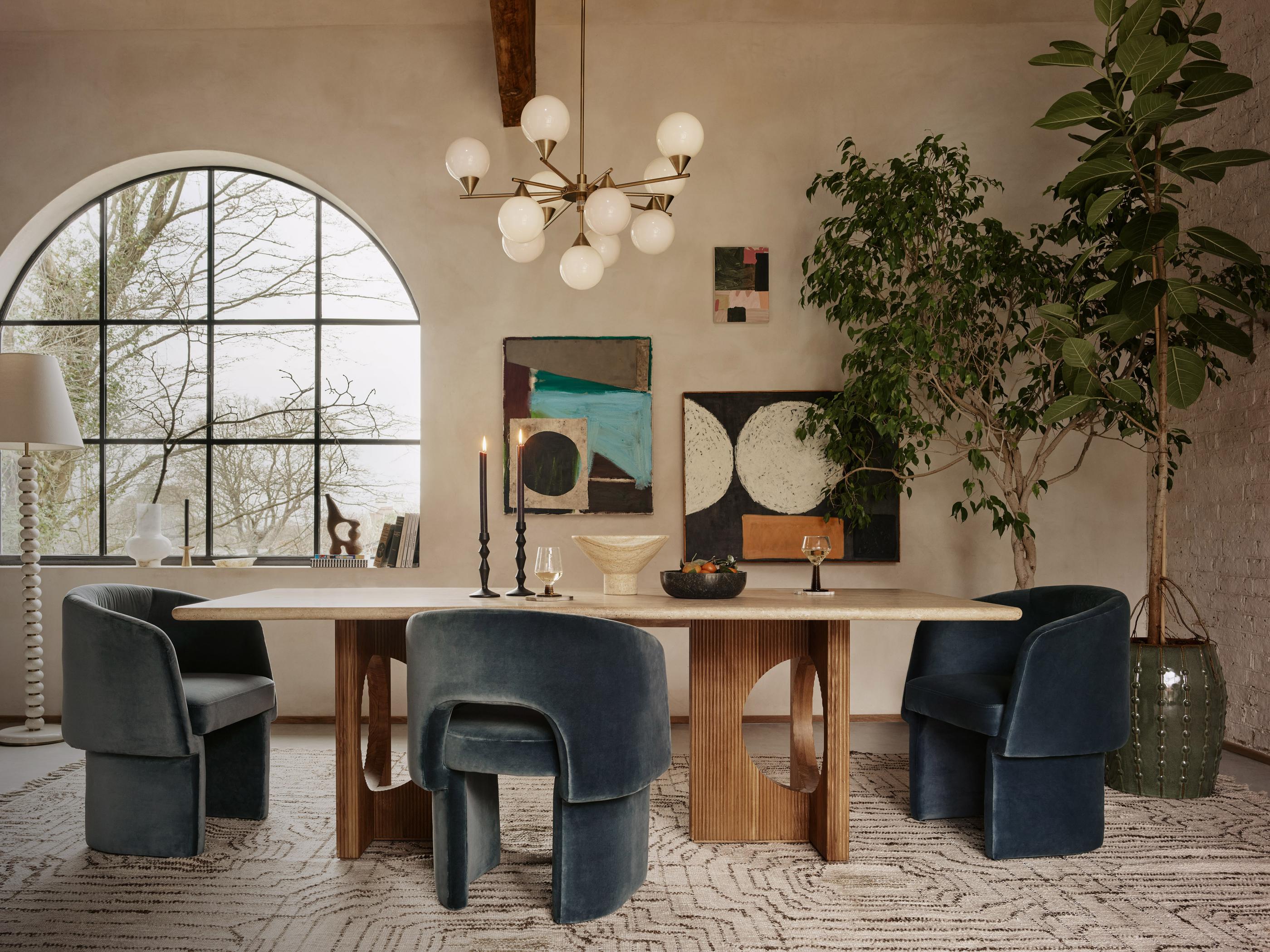 Dining table with blue velvet chairs set for a meal. The room features abstract art, a large arched window, indoor plants, a modern chandelier, and a light-colored rug.
