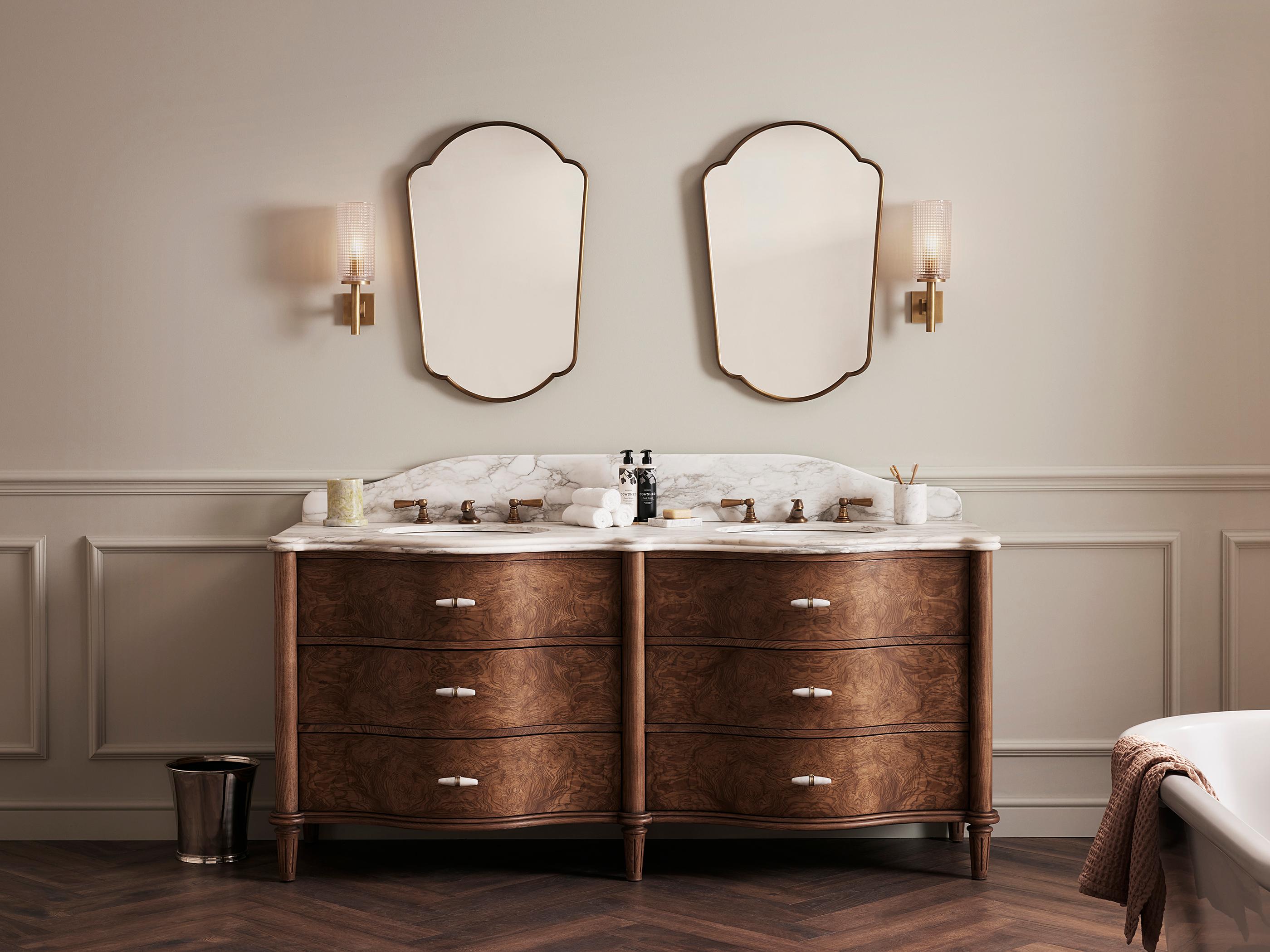 Wooden vanity with marble countertop, twin brass faucets, and two decorative mirrors above. In a classic bathroom setting, adorned with wall sconces, small accessories, and a neatly folded towel.