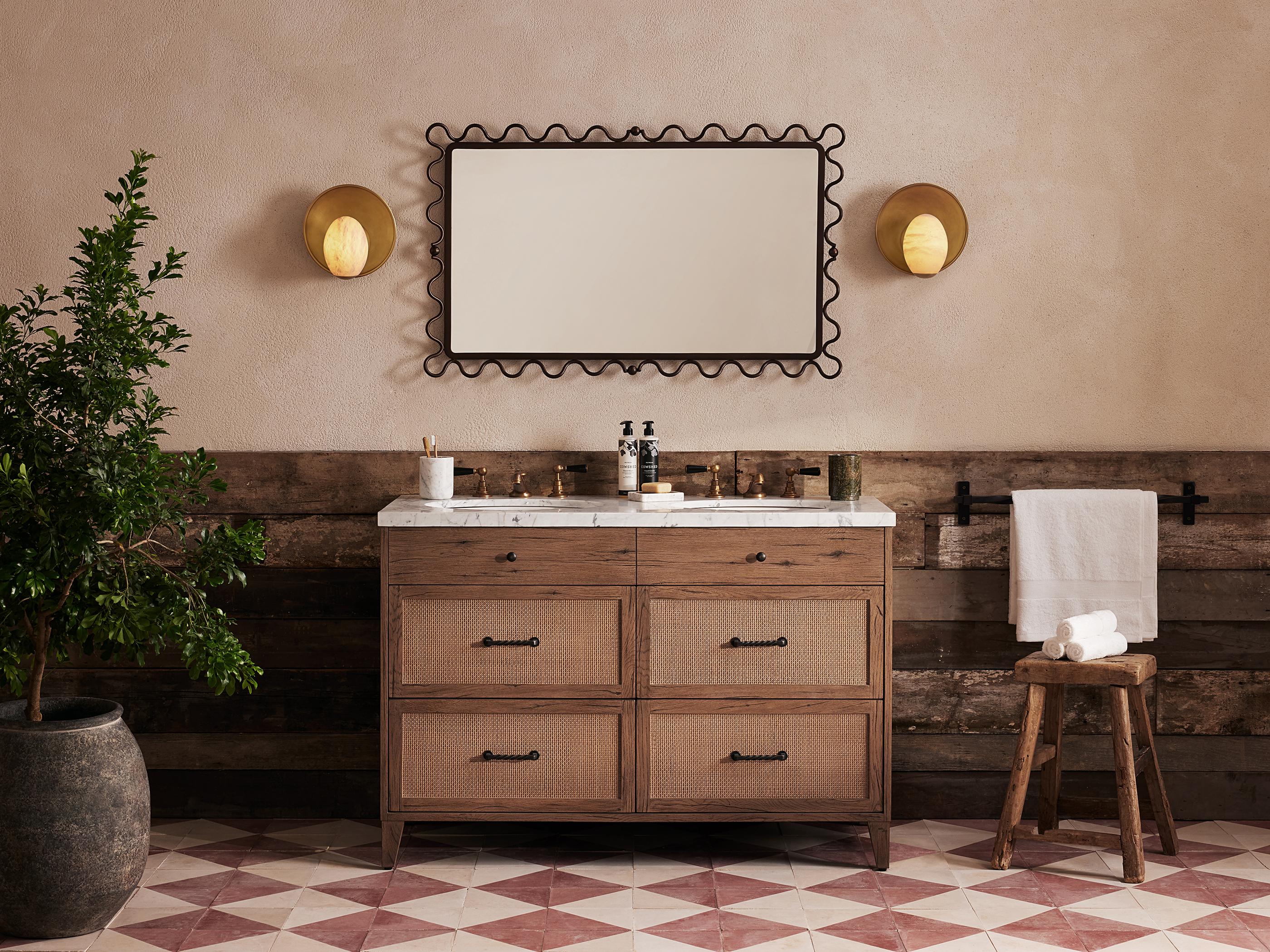 Bathroom vanity with a marble countertop holding toiletries; framed mirror above. Sconce lights flank the mirror. Plant and stool with rolled towels to the sides. Wooden wall paneling and patterned floor tiles.