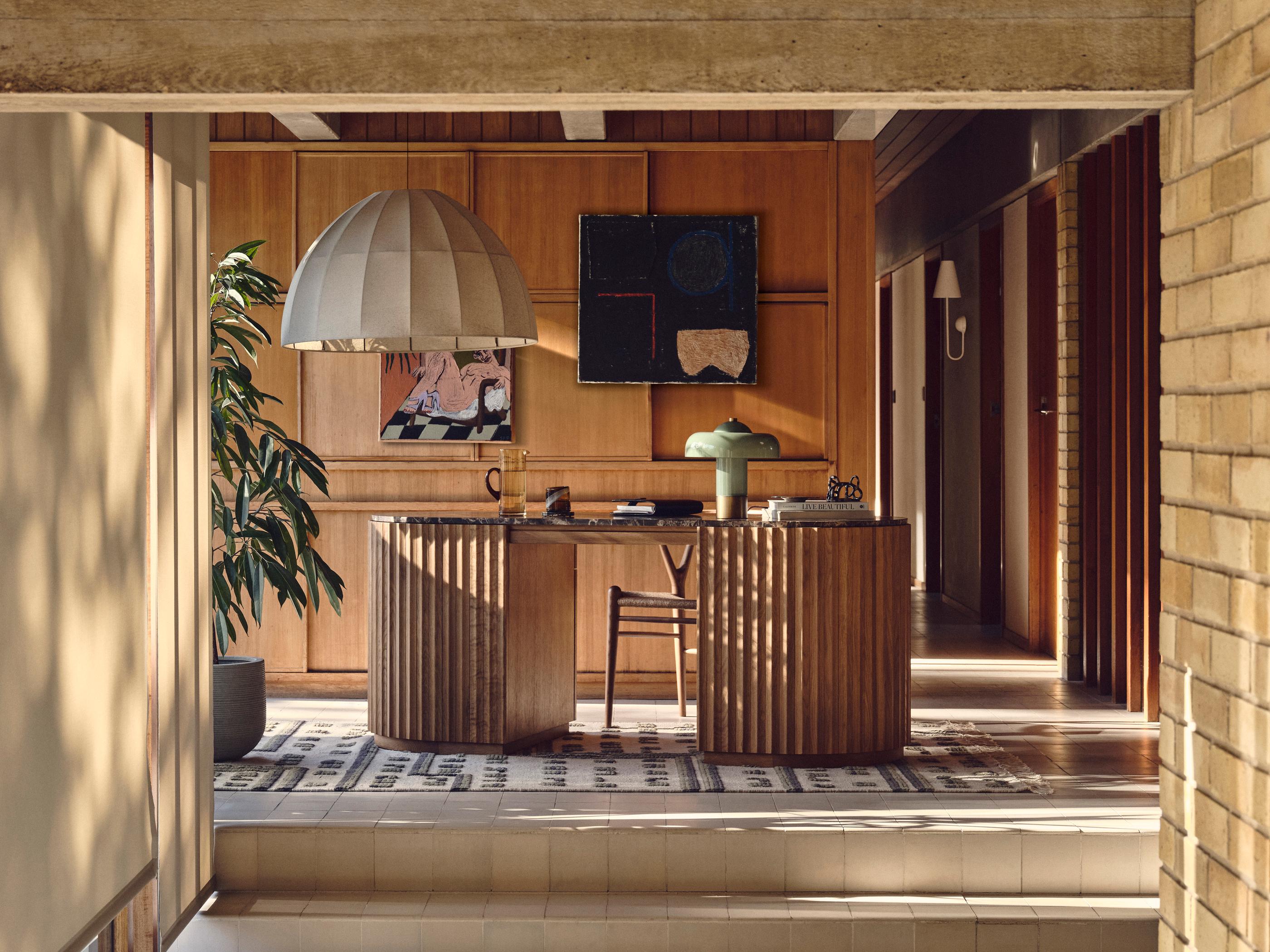 A stylish wooden desk with books and lamp sits in a well-lit room, adorned with a large pendant light, adjacent plant, and modern art on wood-paneled walls.