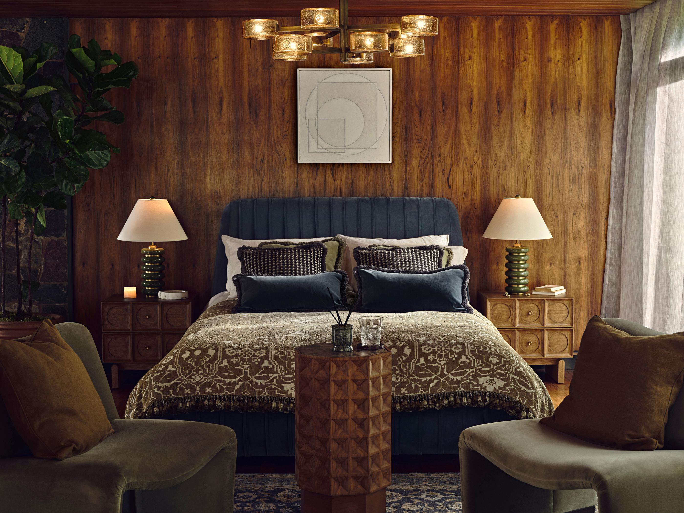 A double bed with patterned bedding and pillows sits against a wood-paneled wall, flanked by two lamps on bedside tables. Two chairs and a small table are in the foreground.