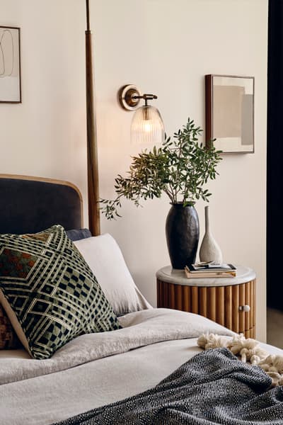 A cozy bedroom features a bed with patterned cushions and a gray throw. A bedside table holds books, dark pottery, and a leafy plant. A wall sconce and abstract art add decor.