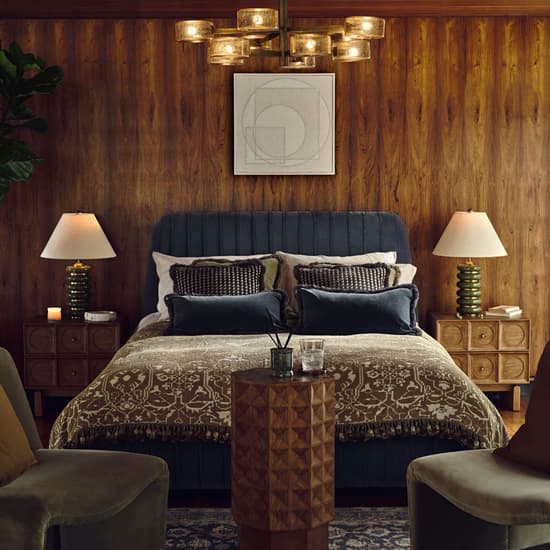 A double bed with patterned bedding and pillows sits against a wood-paneled wall, flanked by two lamps on bedside tables. Two chairs and a small table are in the foreground.