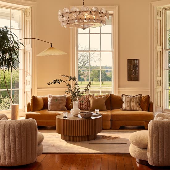 A mustard-colored sectional sofa and two beige armchairs surround a wooden coffee table with vases and glasses in a bright living room, featuring tall windows, a chandelier, and plants.