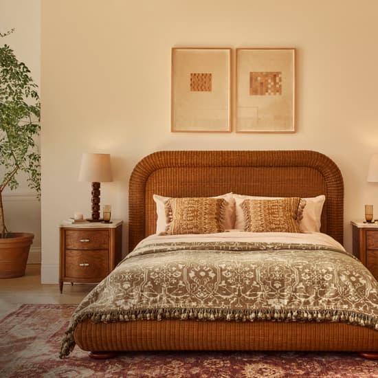 A wicker bed, adorned with patterned pillows and a blanket, is positioned centrally. Flanked by wooden nightstands and lamps, a large potted plant and an armchair decorate the room.
