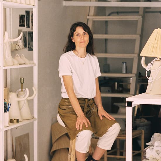 A woman sits on a wooden stool, wearing a white shirt and brown apron, in a cluttered studio with shelves holding various ceramics and art supplies. The background reveals a loft space with a ladder.