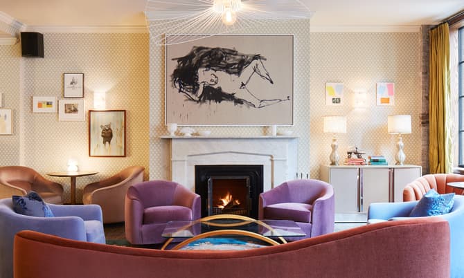 Cozy living room featuring colorful armchairs arranged around a glass coffee table, a lit fireplace with a large abstract painting above it, and art-adorned walls with warm lighting.