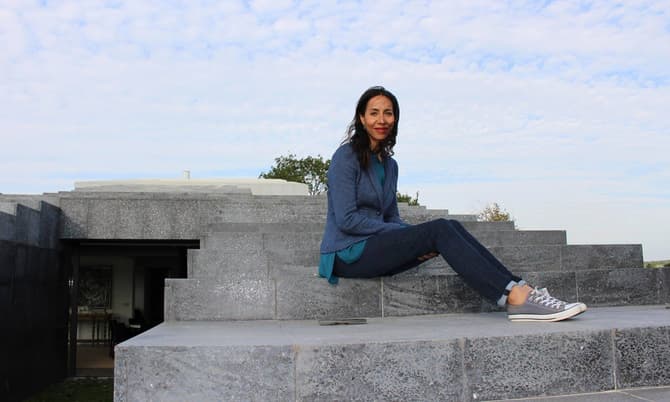 A woman wearing a blue jacket and jeans sits on concrete steps of a modern, grey stone structure, with a partly cloudy sky in the background.