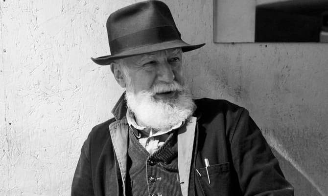 Bearded man wearing a hat and jacket sits against a textured wall, smiling slightly, with a pen visible in his jacket pocket in a dimly lit room.