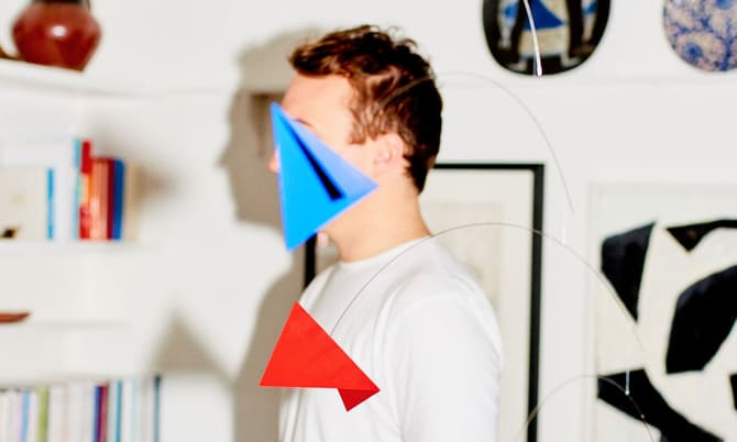 A person stands indoors, partially obscured by geometric shapes (blue and red triangles), with a background of bookshelves and framed artworks on the walls.