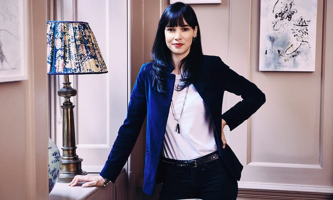 A woman in a white shirt and blue blazer stands confidently with one hand on her hip, indoors by a table with a decorative lamp and framed artwork on the walls.