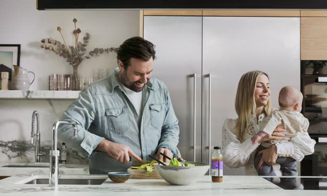 Man preparing a salad in a modern kitchen while smiling, with a woman holding a baby nearby. The kitchen features a marble countertop, sink, and stainless steel appliances.