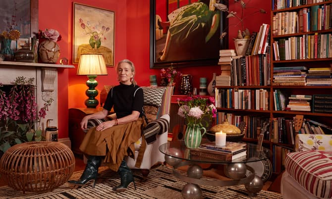 A woman sits on a patterned sofa in a vibrant red-walled living room surrounded by bookshelves, art, flowers, a green lamp, and decorative objects. A striped rug covers the floor.