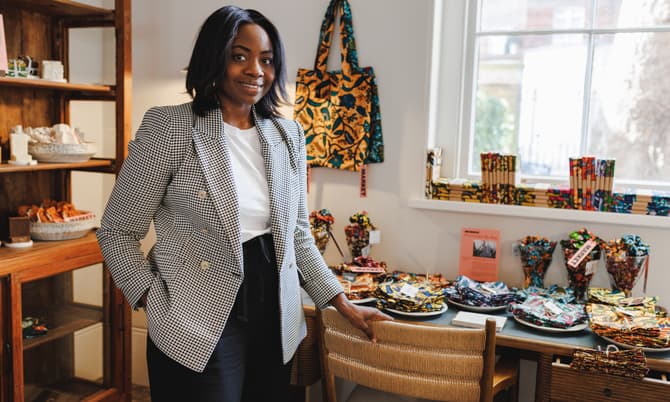 A person wearing a checkered blazer stands beside a table displaying colorful fabrics. The setting is a well-lit room with shelves containing various items and a window in the background.