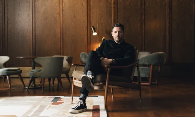 A man sits cross-legged in an armchair, wearing casual clothing and sneakers, in a mid-century modern wood-paneled room with stylish furniture and a patterned rug.