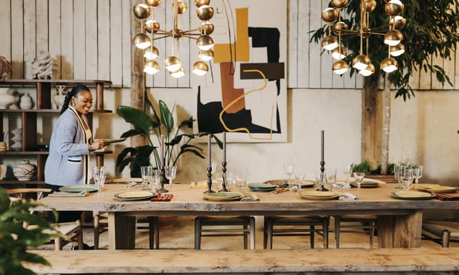 A woman in a light blue sweater is setting a dining table with plates and glassware in a rustic, well-decorated room with plants, shelves, abstract art, and gold chandeliers.