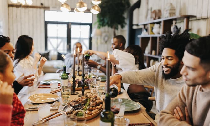 People are sitting around a wooden dining table, eating, drinking, and conversing. Two individuals are pouring wine, surrounded by plates of food, candles, and modern lighting in a cozy, well-decorated room.