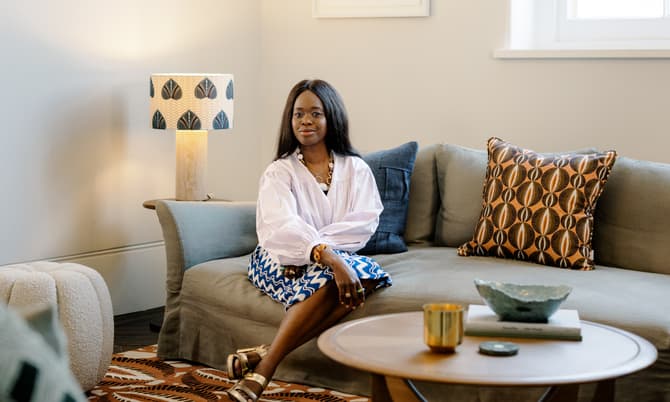 Woman sitting on a grey sofa in a cozy living room, wearing a white blouse and blue patterned skirt. The room features a patterned lampshade, decorative pillows, and a wooden coffee table with books and a candle.