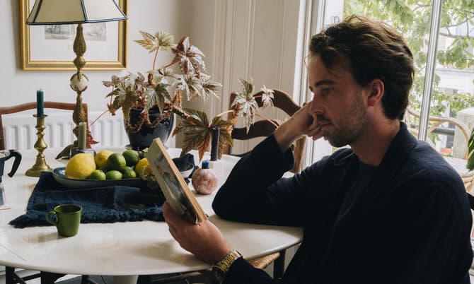 A man seated at a round white table reads a book; the table holds a lamp, plant, and a bowl of fruit beneath framed artworks and beside a large window.