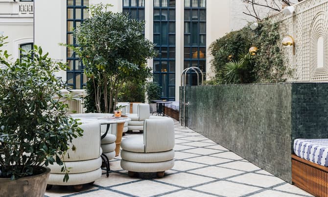 Plush seats surround a table with drinks in a stylish outdoor patio adorned with potted plants and framed by tall windows and a green-tiled wall.