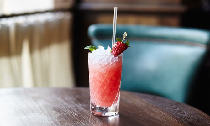A red cocktail with ice, a straw, and a garnished strawberry sits on a wooden table in front of a blurred, elegant indoor setting.