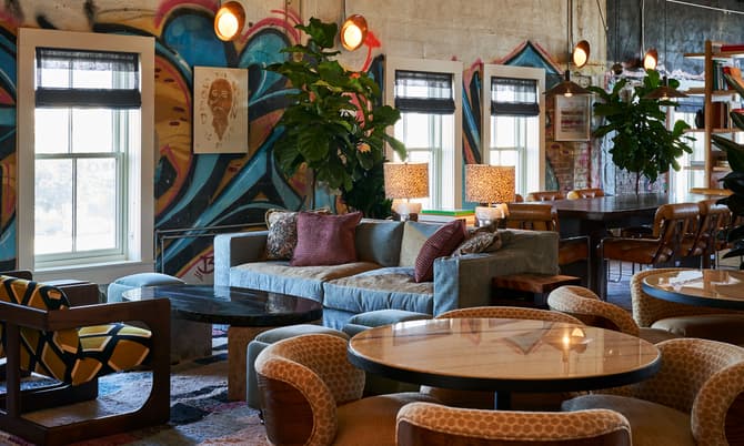 A cozy seating area with a gray sofa, patterned chairs, and round tables is in a room featuring large windows, colorful graffiti walls, hanging lights, and indoor plants.