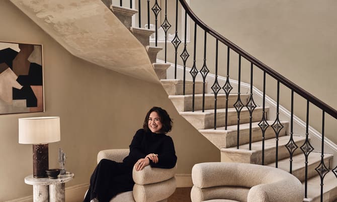 A person in black clothing sits smiling on a cushioned armchair beside a lamp table, under a staircase with an ornate railing. The room features a rug and a minimalist decor.
