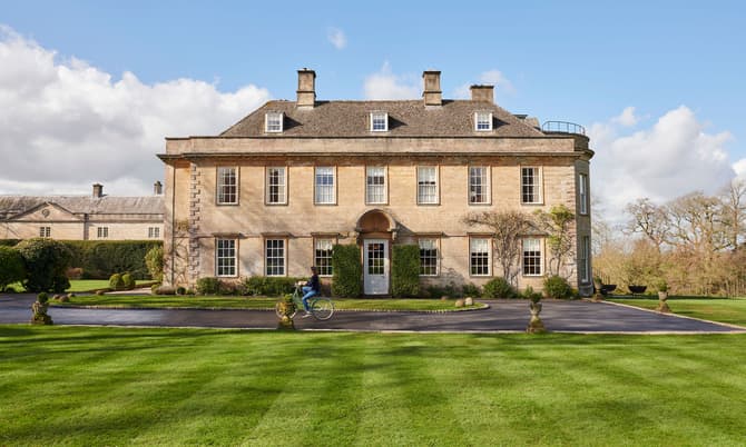 A person rides a bicycle past a large, symmetrical, three-story stone mansion with multiple windows in a landscaped garden with neatly trimmed grass and hedges under a bright blue sky.