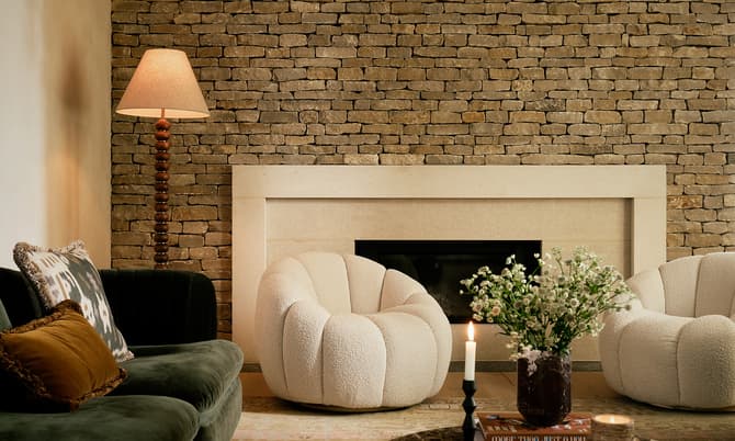 A living room features a green velvet sofa with cushions, two white plush armchairs, a stone fireplace, a standing lamp, and a marble coffee table with a candle, books, and a vase of flowers.