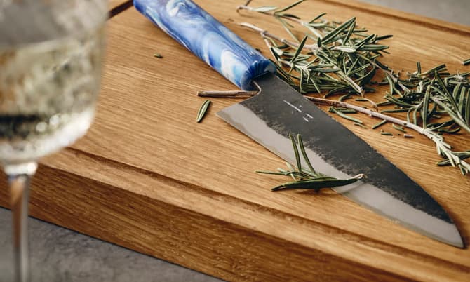A knife with a blue handle lies on a wooden cutting board surrounded by fresh rosemary, next to a glass of white wine and a salt rock on a marble coaster.