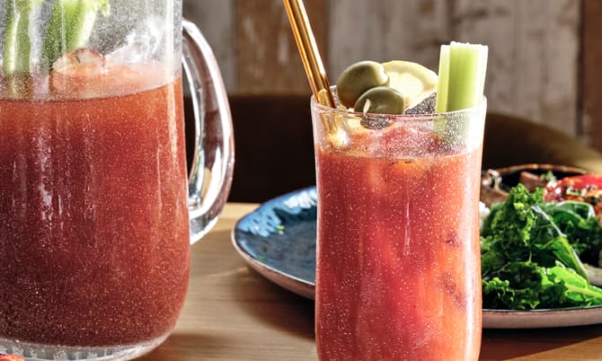 A pitcher and glass of red beverage, garnished with celery and olives, are on a wooden table surrounded by a salad, berries, and a bowl of salt.