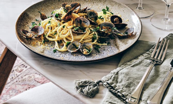 A ceramic plate with pasta and clams sits on a marble dining table with a cloth napkin, silverware, wine glasses, and a bowl of green vegetables in the background.