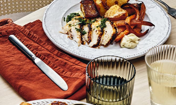 A plate of roasted chicken, potatoes, carrots, and aioli sits on a wooden table set with utensils, a cloth napkin, and glasses of drink, in a cozy dining setting.