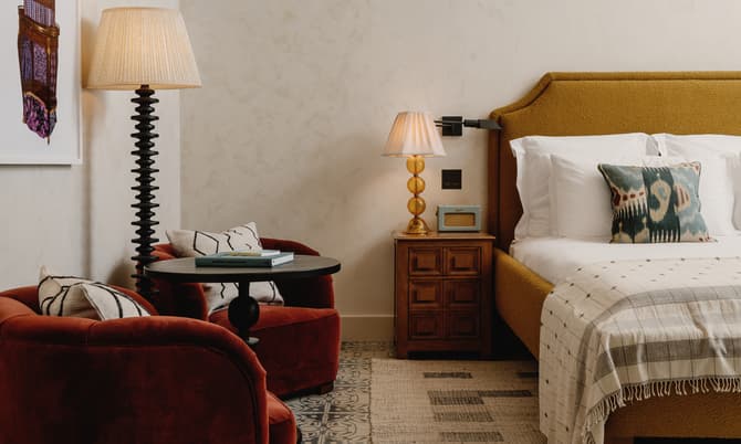 A cozy bedroom features a mustard yellow bed with white linens and a patterned pillow, flanked by a wooden nightstand with a lamp and radio. Two red armchairs, a round table with books, and a floor lamp complete the setting.