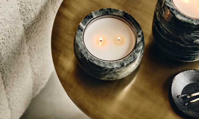 Candles are burning on a marble tabletop beside a textured, beige fabric sofa; a small black dish with matchsticks sits nearby.