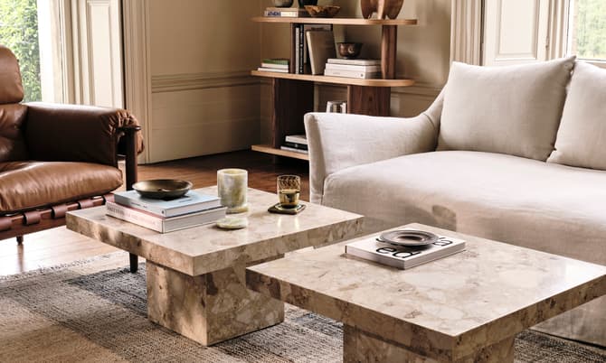 Marble coffee tables displaying books and decor sit between a beige sofa and a brown leather chair; background includes wooden shelves with books and decorative items. Visible book title: "ATHENS RIVIERA".