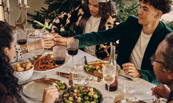 People are seated at a festive dining table, sharing food and drinks, surrounded by decorated Christmas trees and lit candles. Various dishes and wine glasses are spread across a white tablecloth.