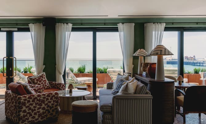 A brightly lit lounge with patterned armchairs, a striped couch, and two table lamps. Floor-to-ceiling windows with sheer curtains overlook an outdoor patio and ocean view.