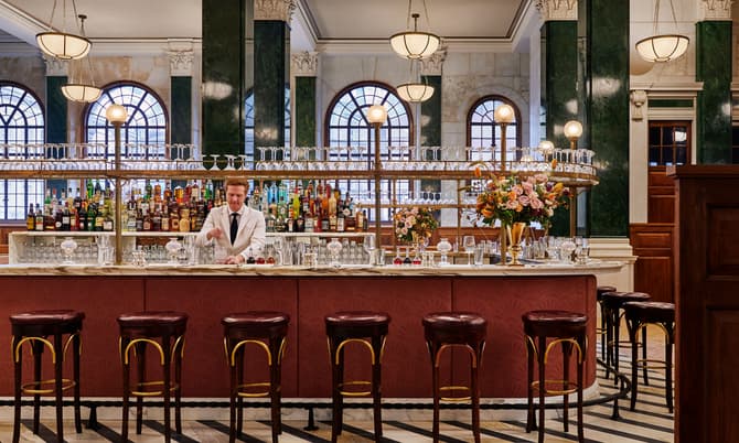 Bartender mixing drinks behind a marble counter, adorned with flower arrangements, in a luxurious bar featuring high ceilings, arched windows, hanging globe lights, and bar stools with dark cushions.